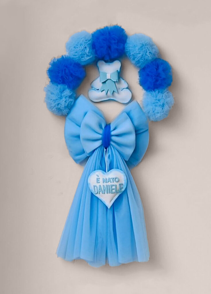 Fiocco bimbo in tulle azzurro con sopra ghirlanda di pompon azzurri e blu e un orsetto pendente; al centro un cuore pendente.