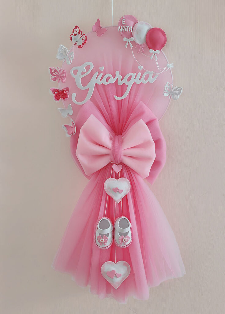 Fiocco nascita bimba in tulle rosa ciliegia con tre pendenti centrali, di cui due cuori e un paio di scarpine neonata e sopra una tela circolare in tulle decorata con impresso il nome "Giorgia".